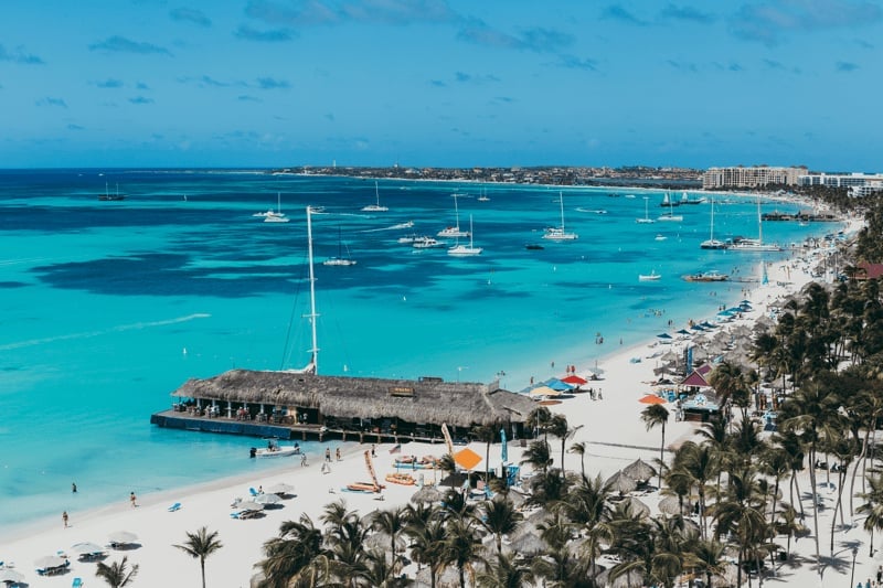 Aerial view of a turquoise Mediterranean beach with sailboats, wooden pier, palm trees, and beachgoers on a sunny day