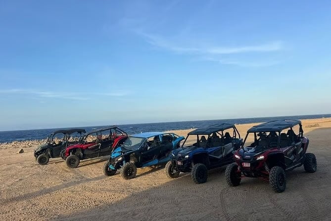 Five off-road buggies lined up on a sandy beach with ocean and blue sky in the background