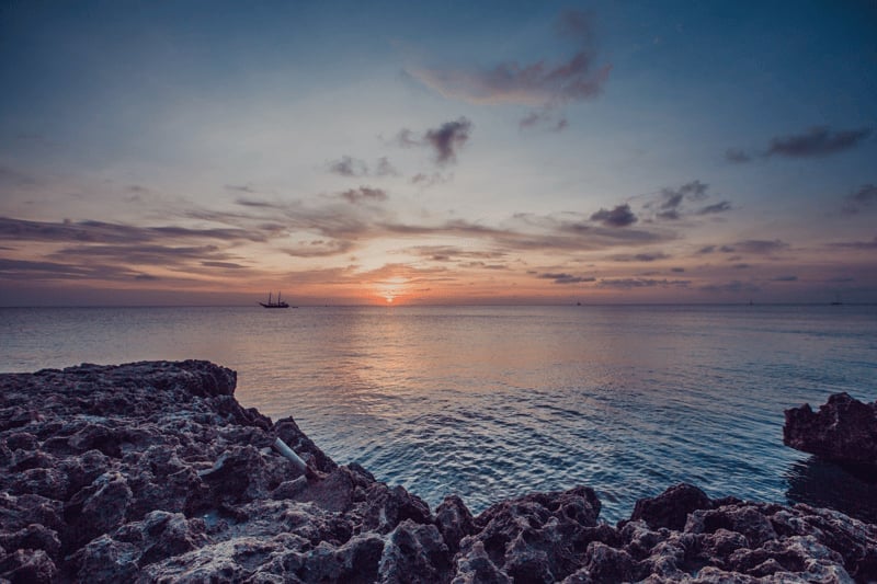 Sunset over calm Caribbean ocean with rocky coastline in the foreground and a distant sailboat on the horizon