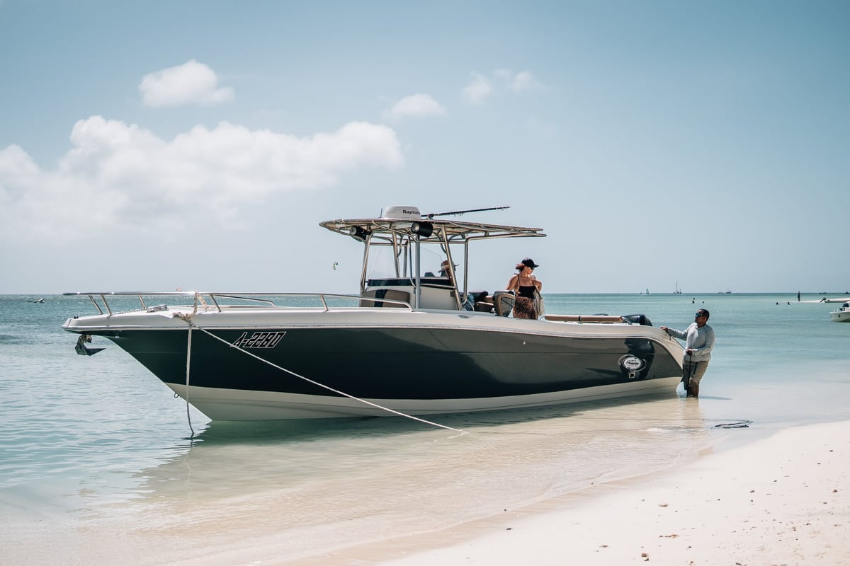 A center console fishing boat moored in shallow turquoise water near a sandy beach with two people visible